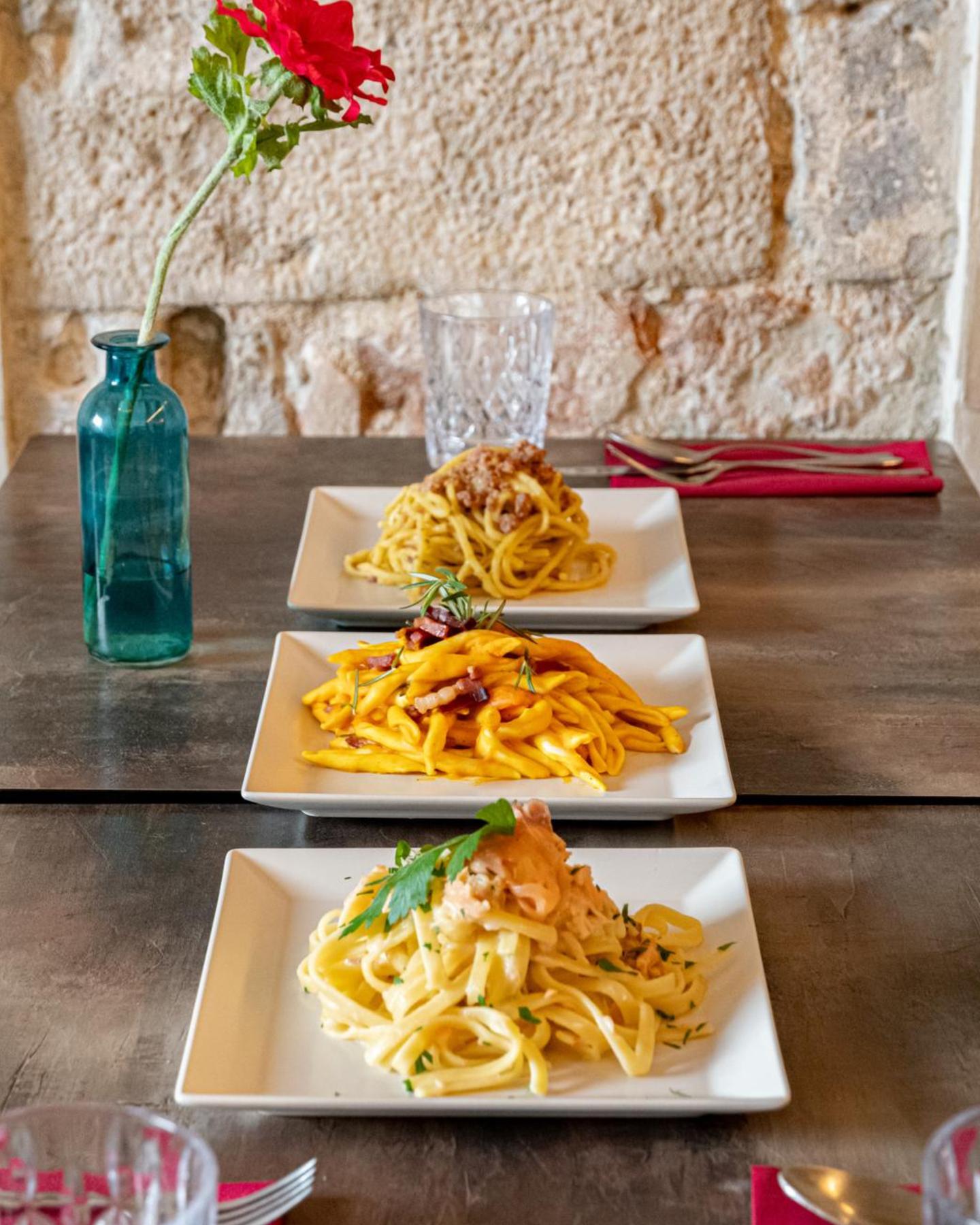 Three pasta dishes in a row on the wooden table in front of the stone wall, with a rose in a vase