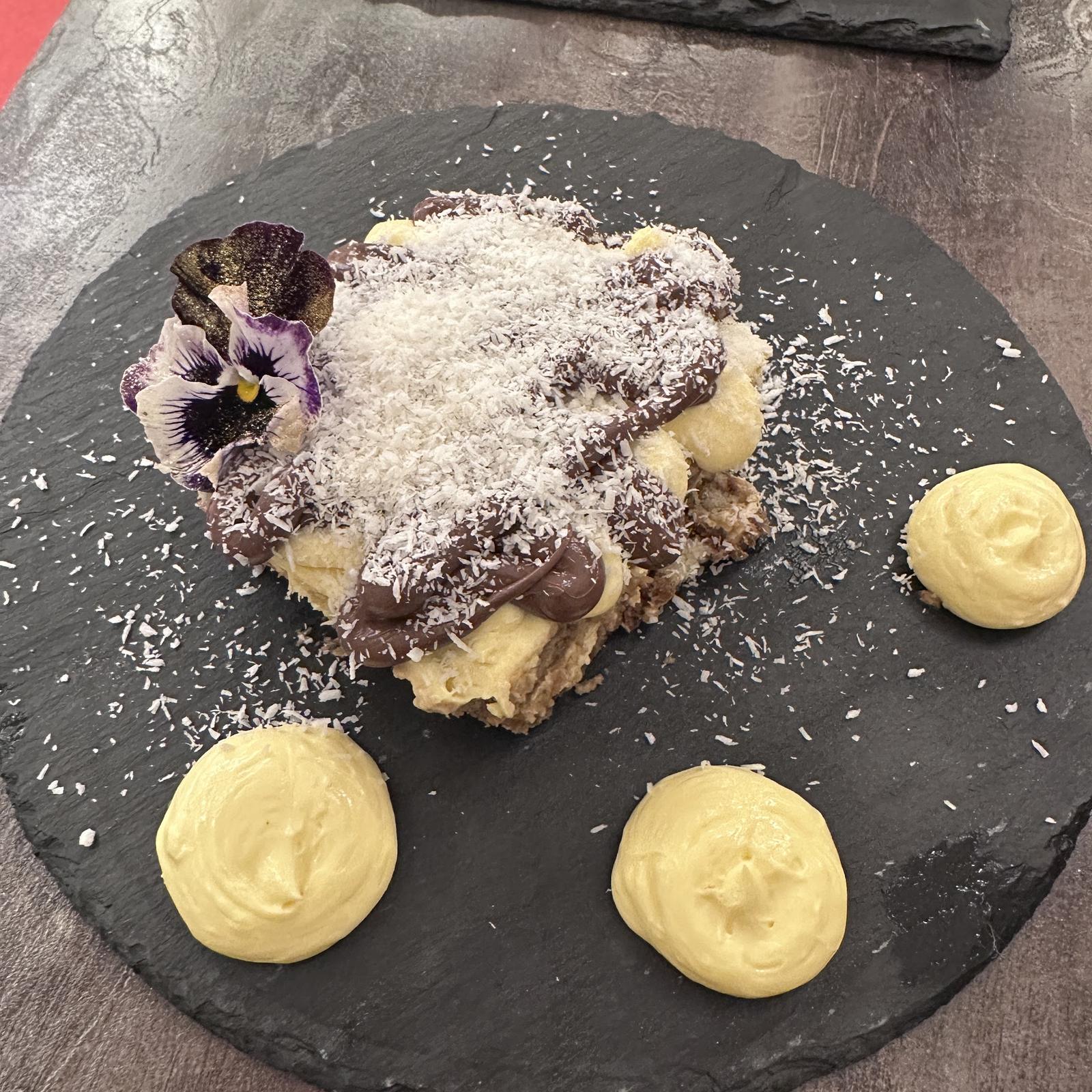 Coconut tiramisù with a pansy and three swirls of cream on a round slate plate